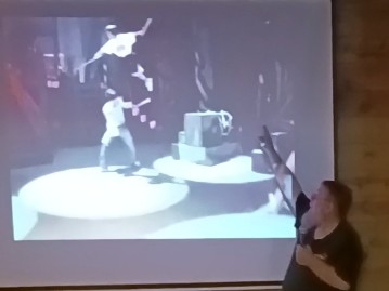 In this screen capture from a short video clip, Tracy Nunnally, MFA, Northern Illinois University (NIU) professor, technical director, head of design and technology, School of Theatre and Dance, points to where the operator is standing in darkness to control the actor’s on-stage flying sword fight. Nunnally, also owner of Vertigo, spoke in the April 17 STEM Café presentation, “Theatrical Special Effects: Engineering Spectacle.” 